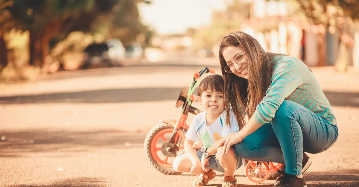 Una madre y su hijo sonriendo con un triciclo en una calle soleada, capturando momentos alegres de unión.