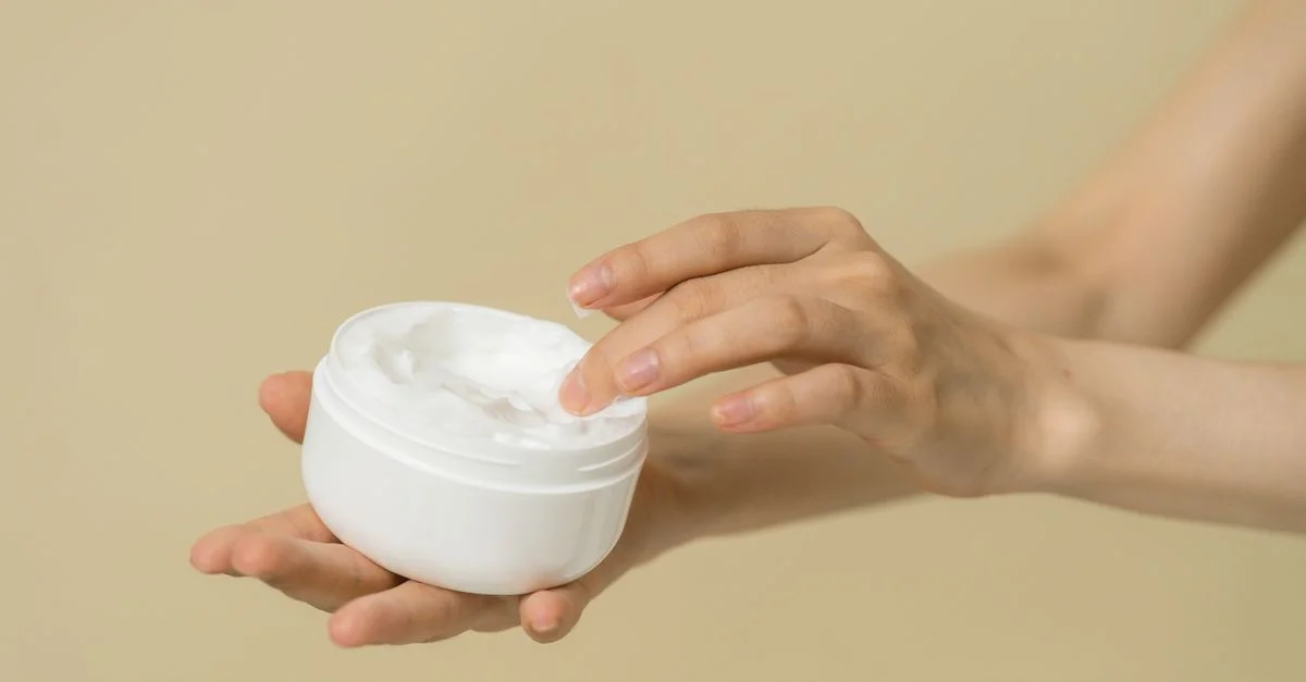 Gros plan sur des mains féminines appliquant une crème hydratante d'un pot ouvert, mettant en avant les soins de la peau et la routine de soins personnels.