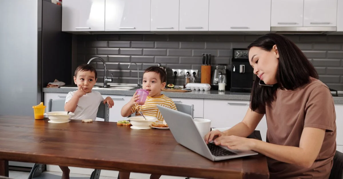 Una madre multitarea con un portátil mientras sus dos hijos pequeños disfrutan del desayuno en la cocina.