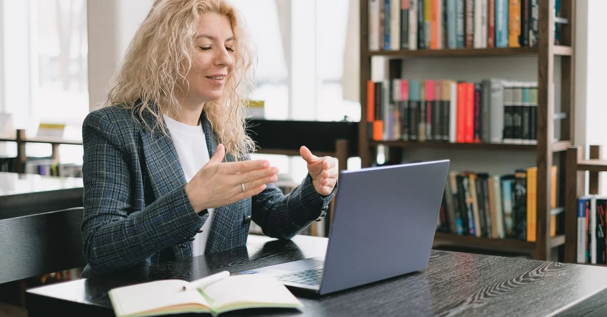 Mujer rubia usando un portátil para videollamada en un entorno de biblioteca, transmitiendo compromiso profesional y educativo.