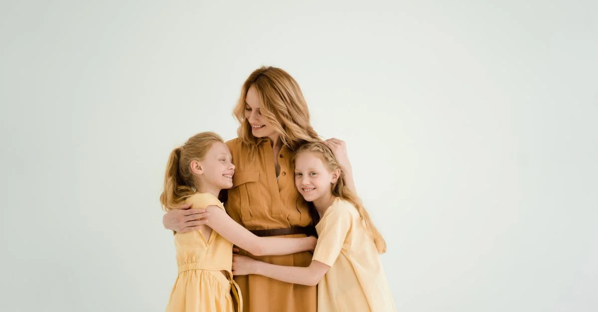 Una madre abraza cariñosamente a sus dos hijas contra un fondo suave y claro, simbolizando el amor familiar y la unión.