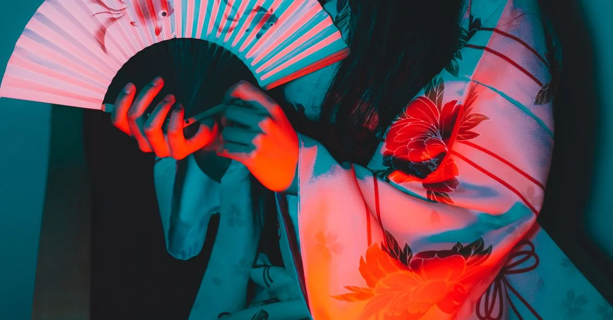 Una mujer en kimono tradicional sosteniendo un abanico, iluminada por luces vibrantes en rojo y azul.